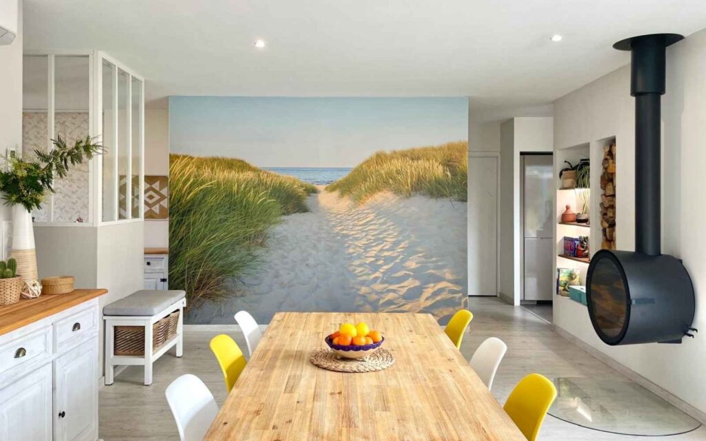 Salle à manger avec papier peint panoramique représentant une plage, bois clair, déco naturelle et cheminée suspendue.